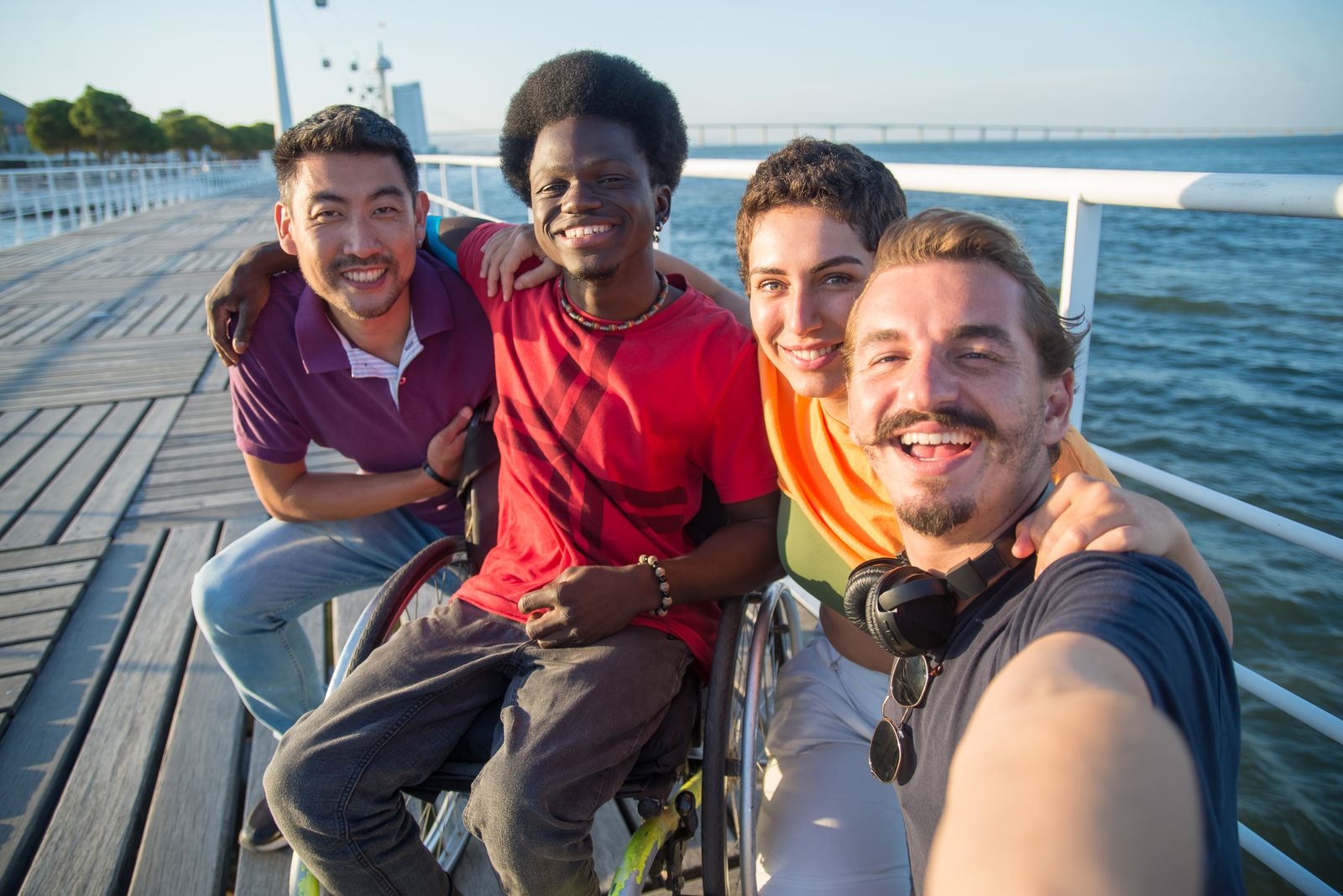 A group of friends, including a young man using a wheelchair, laughing together on a seaside boardwalk
