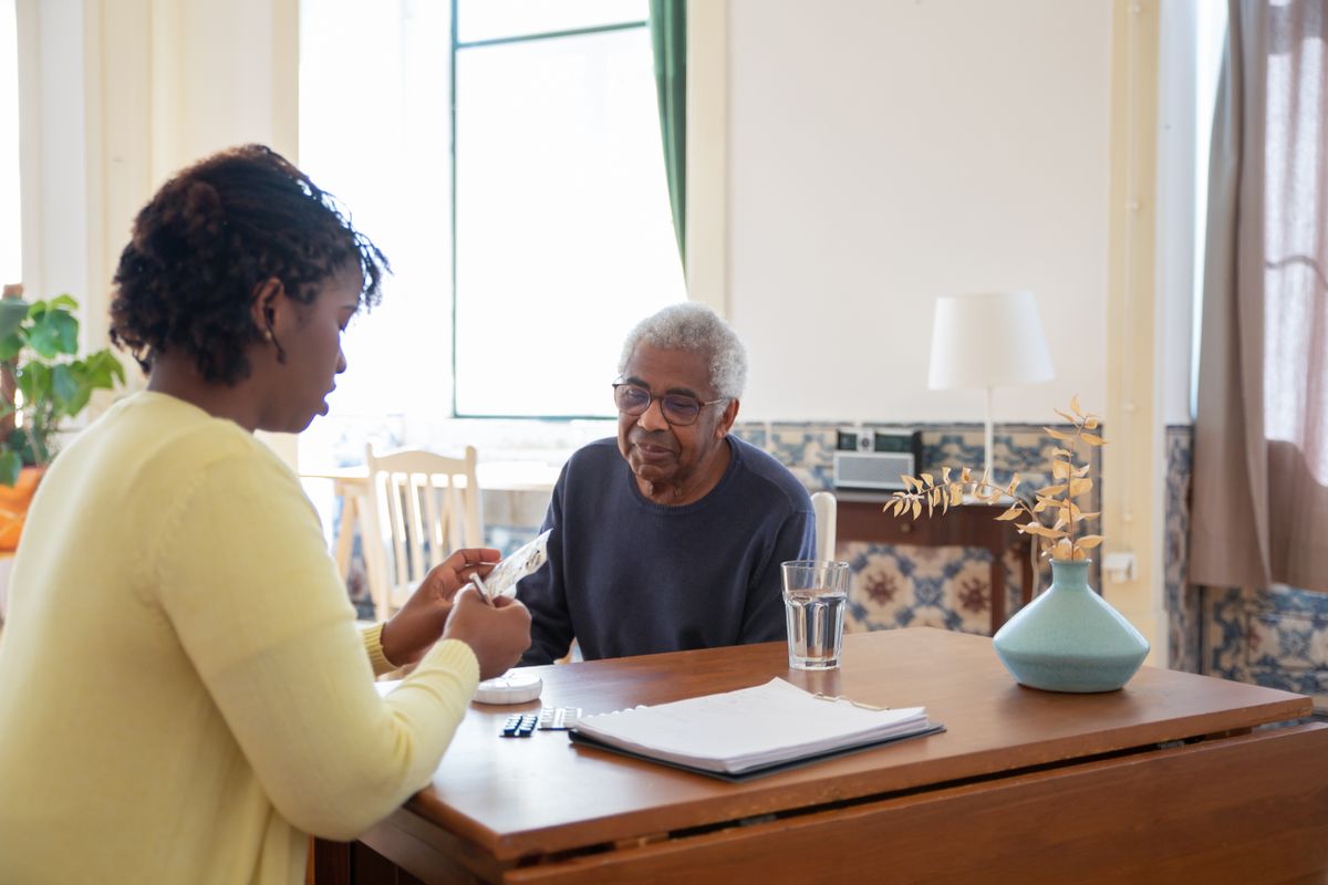 A caring support worker helping an elderly man with medication in a warm, sunlit home