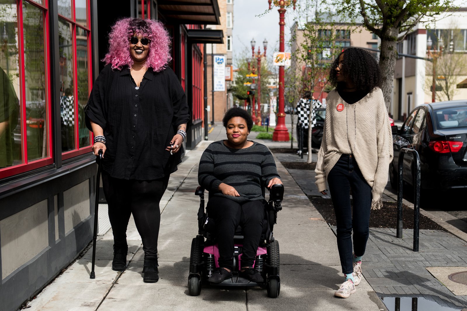 Three young disabled friends walking together down a sunny city street, one using a wheelchair and one with a cane